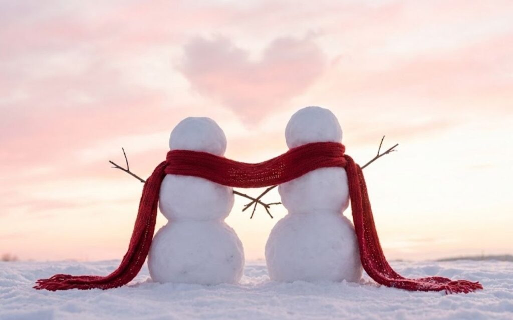 An image of two snowmen standing close together in a snowy field, holding stick hands and sharing a single long red scarf. The clouds in the pink and white sky above them are shaped like a heart.