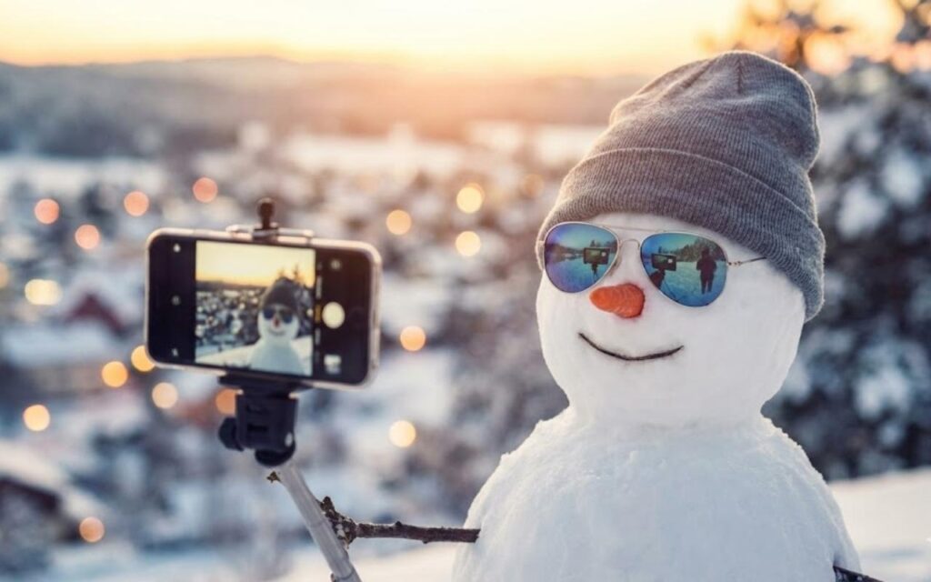 A close up of a "cool" and trendy snowman holding a smartphone on a stick arm to take a selfie. The snowman is wearing sunglasses and a beanie hat, set against a blurred, bright white snowy background.