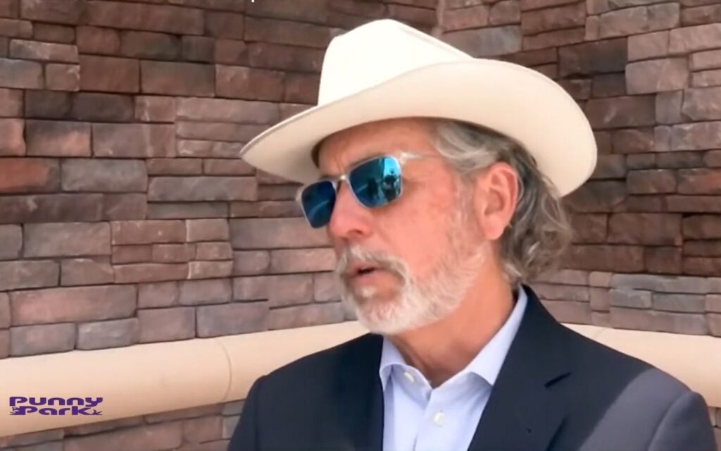 Arch Aplin wearing a white cowboy hat, blue sunglasses, and a suit jacket. He is sitting in front of a wall made of reddish-brown stones.