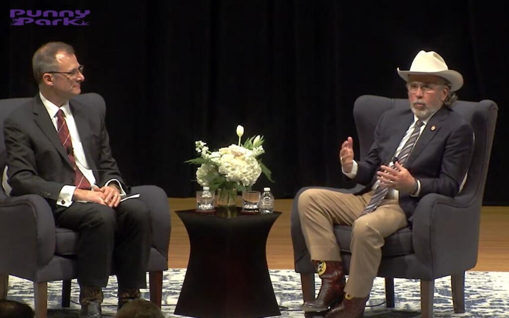 Arch Aplin siting with one other man in armchairs on a stage. One wears a suit, and the other wears a cowboy hat. A small table with white flowers stands between them.